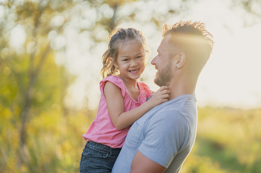 A young Father holds his daughter up in his arms during a walk at sunset on a warm fall evening.  They are both dressed casually and are smiling at one another as they pause to giggle and laugh with one another.