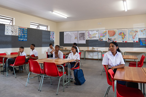 A group of young black children from a rural school in South Africa sit in class listening to their teacher teaching. Children listen to their classroom teacher