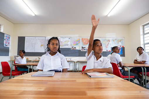 A young African Schoolboy sitting in class at his desk raising his hand eager to answer the teacher's question. Question and answer session as part of the school lesson