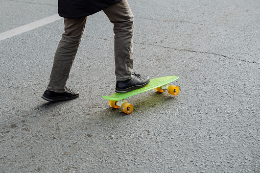 A man in a coat and boots tries to stand on a bright green with yellow wheels pennyboard, skateboard. A hipster learns to skateboard on the asphalt. Activities and new entertainment. High quality photo