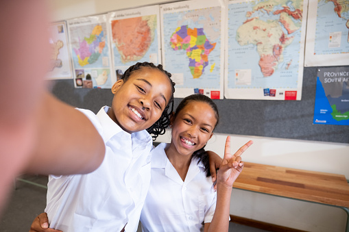 Two African schoolgirls stand in their classroom and pose for a fun selfie photograph. Besties dressed in school uniform taking a photograph.