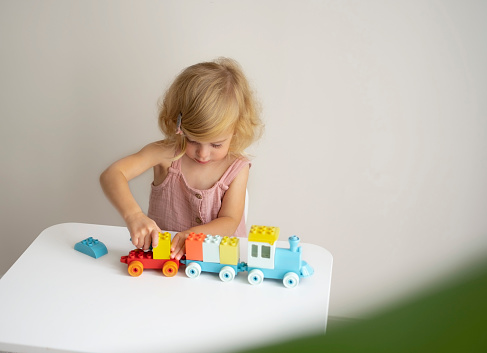 Pretty caucasian 1,2 year old with blond curly hair playing with colourful construction cubes, toys making train.Happy smiling child having fun, early development concept.Copy space