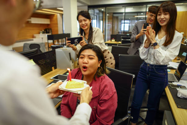 азиатский мужчина, белый воротничок, вошел в офис с небольшой тарелкой торта и отдал ее коллеге, у которого сегодня день рождения. коллега с� - office fun group of people white collar worker стоковые фото и изображения