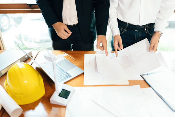 istock Two male engineers look at a project and point to a blueprint about architectural design of an industrial building structure. On the work desk there is a laptop and a wind turbine model in the office. 1785176824