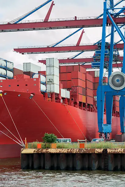 Large red containership docked at the pier of a container terminal.