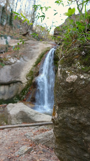 vertical image of a small waterfall in blurred focus visible from behind a rocky ledge, natural landscape for hiking and tourism in the mountains of the Republic of Adygea