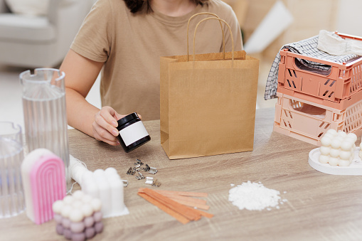 Woman packing homemade candle into paper bag sits by the table