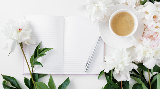 Top view of morning coffee cup, diary with copy space and white peonies flowers on white table