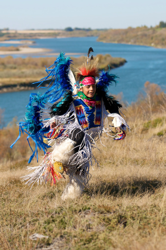 A First Nations boy powwow dancing in a field with a river in the background