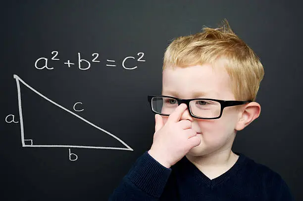 Smart young boy wearing a navy blue jumper and glasses stood infront of a blackboard with the Pythagoras rule explained drawn in chalk