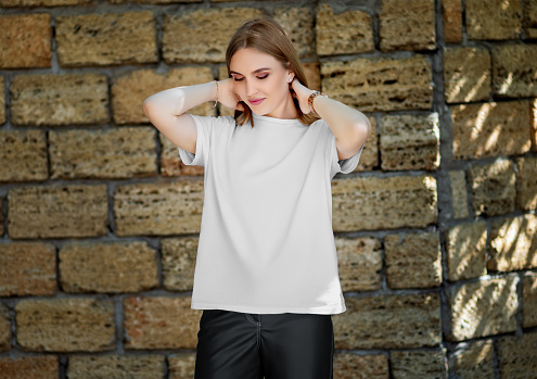 Template of a white t-shirt on a girl, correcting her hair, on the background of a brick wall with shadows, front view. Product photography. Mockup of a women's shirt, clothes for design.