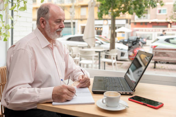 un anciano sentado en una mesa con su ordenador portátil al frente comprobando los datos y tomando notas en su libreta, taza de café y teléfono móvil a su lado - pensionistas trabajadores fotografías e imágenes de stock