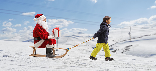 Boy pulling a sleigh with Santa claus sitting and holding a present on a snowy mountain hill
