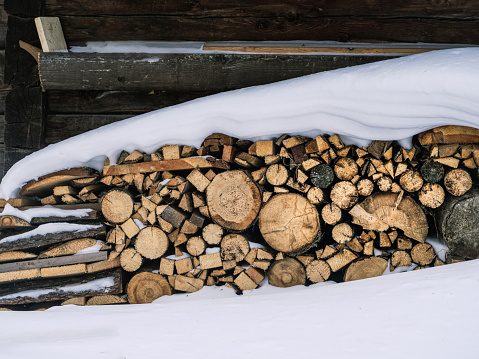 Pile of chopped firewood stock under deep snow next to a wooden building, copy space, winter preparation