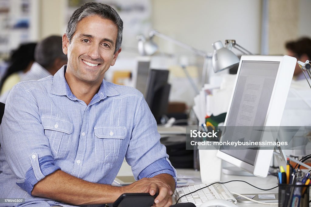 Man Working At Desk In Busy Creative Office Man Working At Desk In Busy Creative Office Looking At Camera Smiling Men Stock Photo