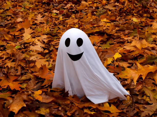 A smiling Halloween ghost made of sheets and fabric sits in fallen autumn leaves.