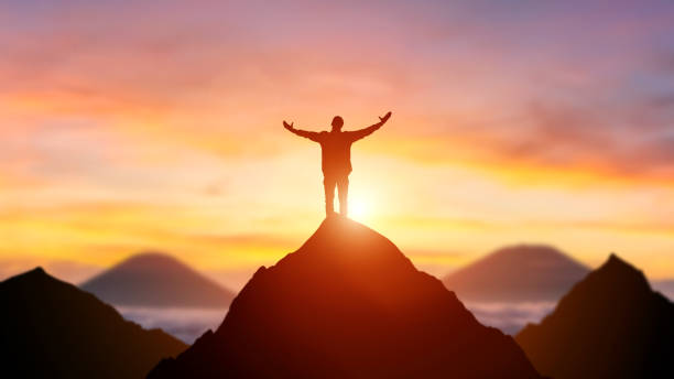 Silhouette of a business person on the top of a mountain peak in sunset background. Winner and conquer of businessman concept. stock photo