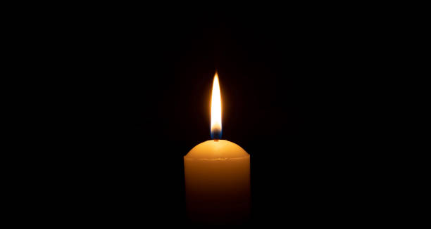 istock Single burning candle flame or light glowing on a big white candle on black or dark background on table in church for Christmas, funeral or memorial service with copy space. 1785513686