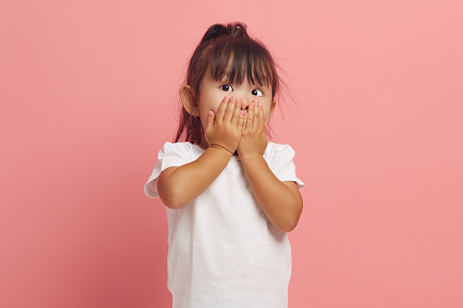 Studio portrait of shocked little girl covers mouth with hands stares scared at camera cannot believe in something horrible dressed in white t shirt isolated over pink background. Omg concept.