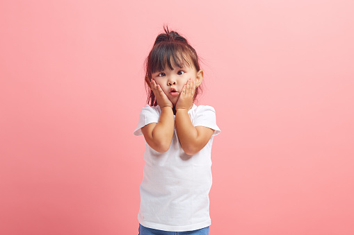 Studio portrait of astonished little girl keeps hands on cheeks stares with bugged eyes, isolated over pink background.