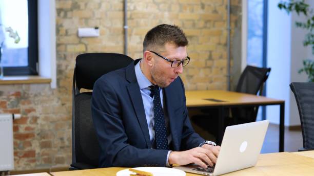 istock Slow motion video. Middle age Caucasian man is sitting in the office, working on computer and he eating and bites big chunks of sandwich. 1785553459