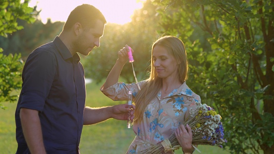 Close up of cheerful cute couple in love together outside on nature. Man and woman with flowers standing in park and blowing soap bubbles. Love and romantic. Boyfriend girlfriend relationships