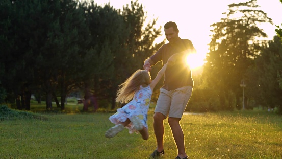 Young Caucasian male parent playing with little daughter on nature outdoors. Father holding child for hands spinning girl in park. Kid having fun with dad at sunset. Summertime. Family leisure concept
