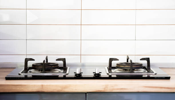 Front view of Contemporary tempered glass gas stove hob with Two burners with auto ignition knob on wooden countertop, cast iron pan supports fan hood and oven built in compact stock photo