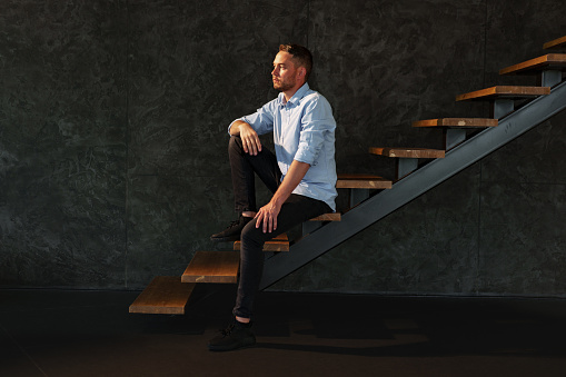 Businessman sitting on a ladder in a dark room, young man in formal wear portrait