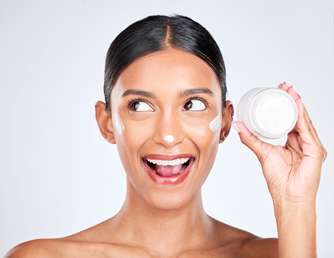 Woman, happy surprise with cream on face and beauty, moisturizer and skincare with container on white background. Cosmetics product, lotion or sunscreen, facial and skin with dermatology in studio