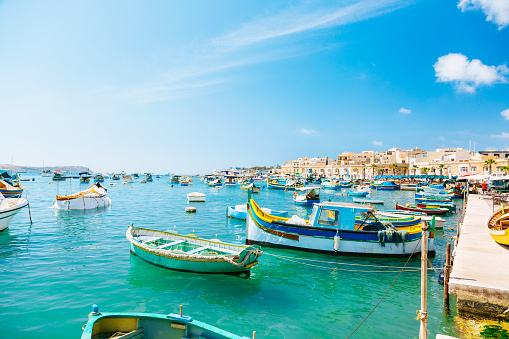 A view of the harbor in Marsaxlokk, Malta, with colorful boats in the foreground and the town’s skyline in the background. The boats are traditional Maltese fishing boats, known as luzzus, and are painted in bright colors with a blue sky and sea in the background. The boats are equipped with a pair of eyes on the bow, which are believed to protect the fishermen from evil spirits.