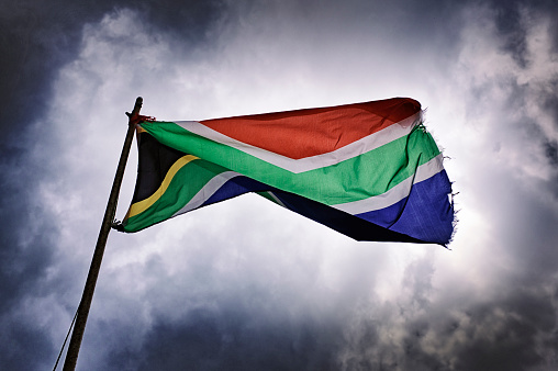The South African flag flies undaunted, its colors vibrant against the brooding sky, a poignant symbol of hope and unity in the face of life's tumultuous challenges.