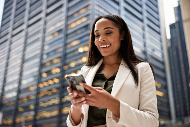 Businesswoman smiling while text messaging good news Close waist up view of longhaired Black professional in late 20s wearing office attire and communicating on portable device, NYC office building in background. people using mobile phone stock pictures, royalty-free photos & images