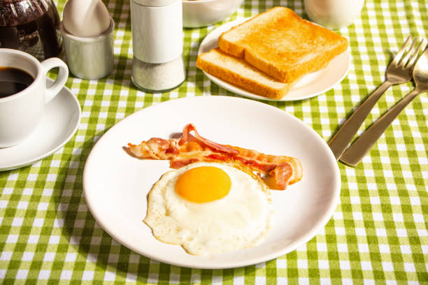 Fried egg with roasted bacon, black coffee, toasts on checkered tablecloth. Retro American breakfast. Hard shadow