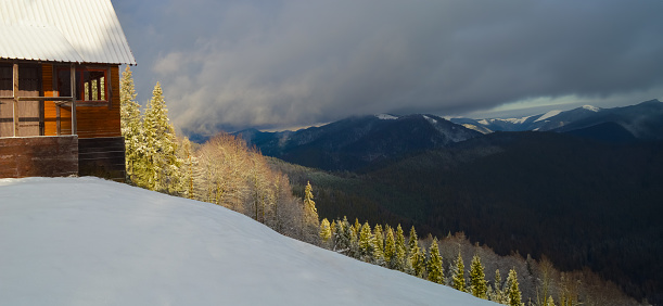 A steep mountainside covered in snow. Coniferous forest illuminated by bright sunlight. Panoramic mountain views and dramatic skies in winter. Part of the wooden house in the foreground.