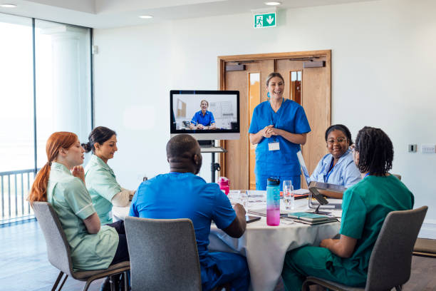 Efficiency and Excellence A group of healthcare professionals sitting around a table, taking part in a medical training meeting in a medical building in Whitley Bay, North East England. They are all listening to a webinar on a computer that a nurse is hosting while they listen to another healthcare professional talking to the whole group as she stands next to the screen.<br><br>Videos available for this scenario. Hospital stock pictures, royalty-free photos & images