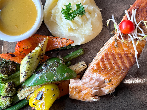 Stock photo showing close-up, elevated view of a healthy meal consisting of a dry fried salmon fillet served with a ramekin of lemon garlic butter sauce, mashed potatoes and roasted vegetables including slices of orange, yellow and green bell pepper, broccoli, baby corn and  green beans. This dish is part of a healthy eating, low calorie diet plan.