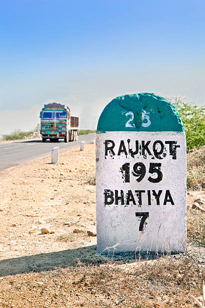 Direction sign and milestone to Rajkot India Dwarka Roadtrip. Vertical take of 195 kilometer milestone and direction sign to Rajkot in Gujarat India with a typical truck about to pass by. Rural country side location in the summer season with dry barren wasteland of the region rajkot india stock pictures, royalty-free photos & images