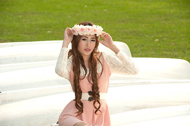 A close-up portrait of a young woman wearing a delicate floral wreath made of pink and white flowers against a white background. Her gentle smile and natural beauty create a soft, romantic, and fresh atmosphere.