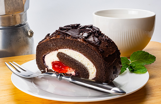 Homemade sponge chocolate roll cake with metal fork and green mint leaves in white ceramic dish and a white coffee cup on wooden board.