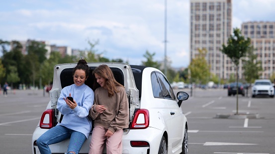 Joyful young adult beautiful woman sitting in car trunk with teen girl kid looking at smartphone screen swiping and tapping on gadget while laughing. Mother and daughter surfing internet on cellphone