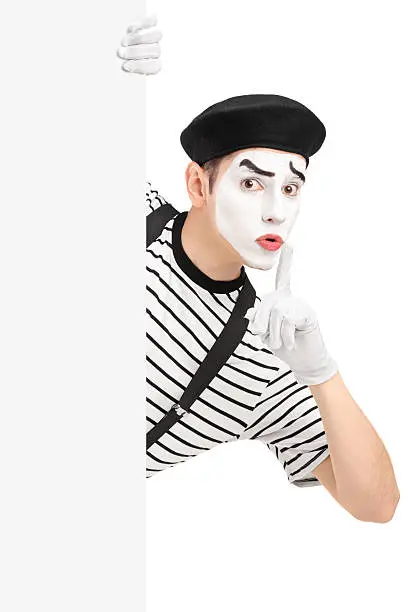 Male mime artist holding a blank panel and gesturing silence with a finger on his mouth, isolated on white background