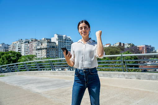 Medium shot front view of Latin-American woman looking at camera while raising a fist and holding smart phone in a park