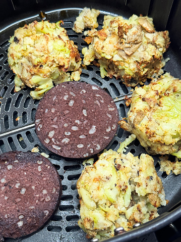 Stock photo showing close-up, elevated view of the inside of an air fryer with bubble and squeak piles (mash potatoes, onions and cabbage) and slices of black pudding being cooked. Healthy cooking concept.