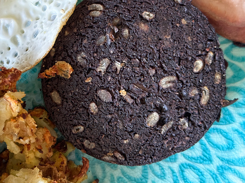 Stock photo showing close-up view of a blue plate containing sunny-side-up fried egg, fried bubble and squeak, black pudding slice and grilled bacon rasher.