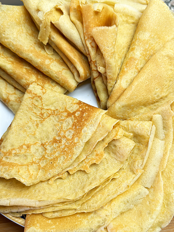 Stock photo showing a close-up, elevated view of a wood grain kitchen counter with a white plate containing a spiral pile of freshly made French crepe pancakes which have been folded into quarters.