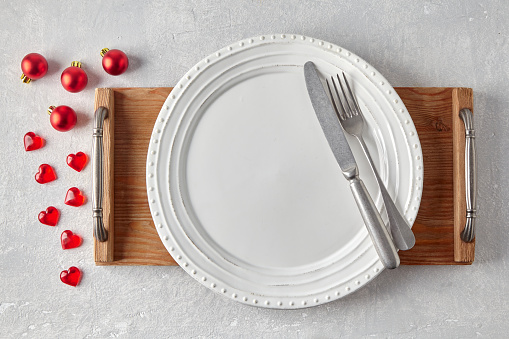 Ceramic white plate with cutlery on a white wooden tray on a gray concrete table surrounded by red Christmas balls and hearts. Layout for displaying food in the New Year's menu. Top view with copy space