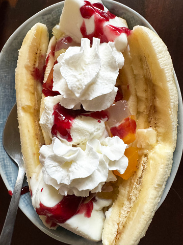 Stock photo showing close-up, elevated view of restaurant dessert of a portion of banana split with fruit salad, vanilla ice cream and aerosol whipped cream in a bowl. This indulgent pudding is served with raspberry sauce.