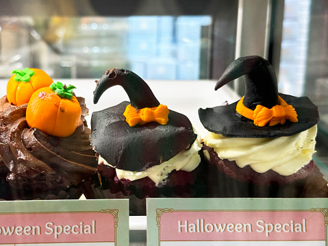 Stock photo showing close-up view of bakery patisserie display showing rows of indulgent Halloween designed desserts on a shelf behind a glass panel.