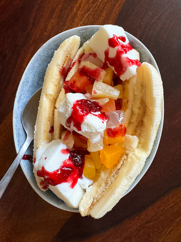 Stock photo showing close-up, elevated view of restaurant dessert of a portion of banana split with fruit salad, vanilla ice cream and aerosol whipped cream in a bowl. This indulgent pudding is served with raspberry sauce.